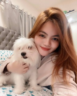A young woman with long light brown hair hugs a small white dog on a bed with patterned sheets, smiling at the camera.