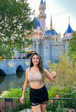 A smiling woman with long hair in a beige crop top and black denim shorts poses in front of a fairytale castle at a