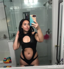 Brunette woman in a black cutout bodysuit takes a mirror selfie in a bathroom, showing her curvy figure.