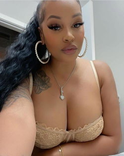 Curvy woman with long dark hair, wearing a beige lace bra and large hoop earrings, posing indoors with visible arm