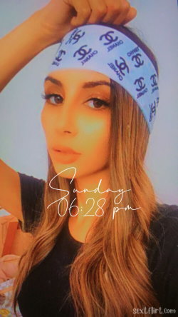 A woman with long brown hair and a patterned headband poses in a selfie, wearing a dark top against a plain background.
