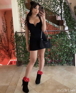 Brunette woman in a black mini dress and black boots with red ruffles takes a mirror selfie in a lush, plant-covered