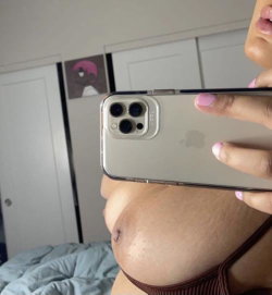 A woman with dark skin takes a mirror selfie in a bedroom, wearing a brown bra, holding a phone with pink nails visible.
