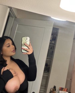 Curvy woman with dark hair takes a mirror selfie in a black top, partially unbuttoned, revealing bare breasts in a