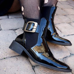Close-up of shiny black patent leather ankle boots with gold buckle, worn with patterned fishnet stockings on a stone