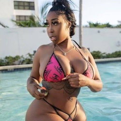 Curvy woman in a pink patterned bikini poses by a pool, holding a small object, with visible tattoos on her torso.