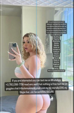 Blonde woman with curly hair takes a mirror selfie in a gray top, showing her backside in a bedroom setting.