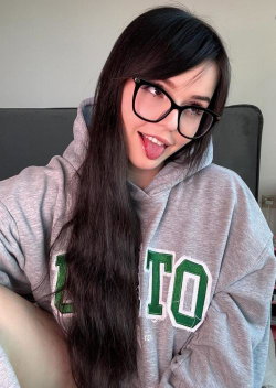 A young woman with long dark hair and black glasses sticks her tongue out playfully while wearing a gray hoodie, seated