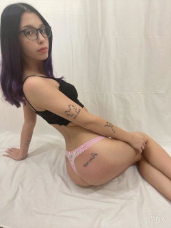 Woman with glasses and purple hair in black bra and pink underwear on a bed.
