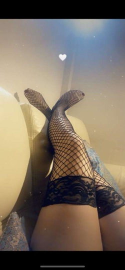 Close-up of legs in black fishnet stockings with lace trim, resting on a light-colored couch with soft lighting and