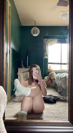 A woman with shoulder-length hair sits nude on the floor in a bedroom, taking a mirror selfie while wearing fluffy