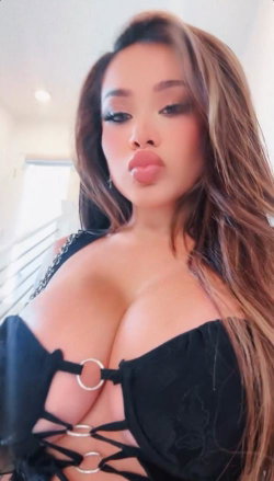 Asian woman with long brown hair wearing a black lace-up top, making a playful face indoors.