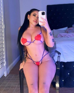 Curvy woman with long dark hair takes a mirror selfie in a red heart-themed lingerie set, sitting on a chair in a