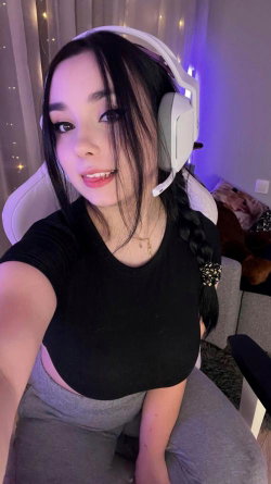 A young woman with long dark hair in a braid wears a black crop top and gray pants, sitting in a gaming chair with