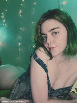 A woman with short green hair lies on her side in a dark lace top, surrounded by string lights against a teal backdrop.