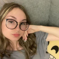 A woman with wavy blonde-brown hair and cat-eye glasses lies on a gray couch, resting her head on her hand in a casual