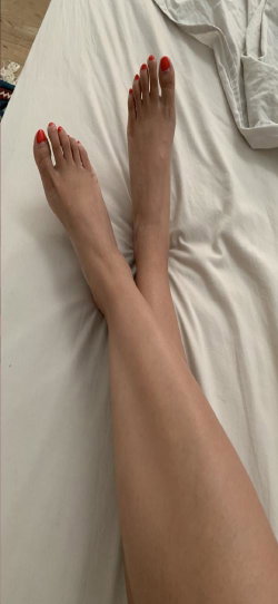 Close-up of bare legs and feet with red toenails resting on a white bedsheet, suggesting a relaxed indoor setting.