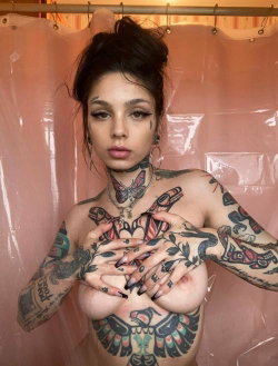 A tattooed woman with dark hair in a bun covers her bare breasts with her hands, standing in front of a pink shower