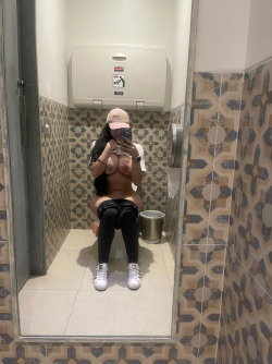 A woman with long dark hair sits on a toilet in a tiled bathroom, topless and wearing black pants and white sneakers,