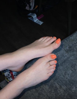Close-up of feet with bright orange toenail polish, resting on a gray textured surface, one foot adorned with a small