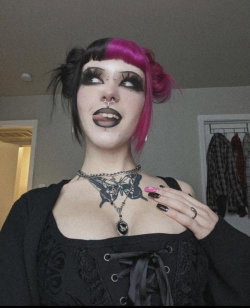 Person with black and pink hair, dark makeup, nose ring, and neck tattoo wearing a black corset-style top indoors.