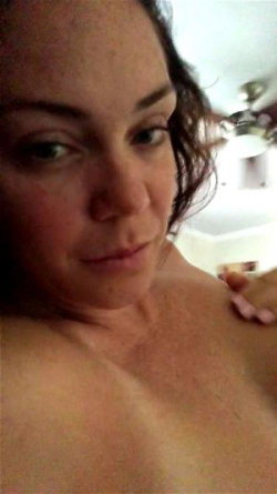 Close-up selfie of a woman with dark hair, bare shoulders, and visible chest, indoors under warm lighting.