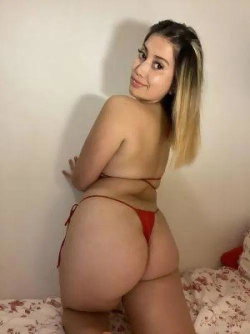 Curvy woman with ombre hair posing in red thong lingerie on a bed, back to camera with head turned.