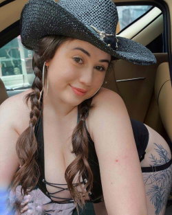 Curvy woman with braided brown hair and a black cowboy hat, wearing a strappy top and showing a tattooed thigh, seated