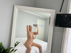 Blonde woman with curvy build takes mirror selfie nude on white bed, holding phone in one hand, arm raised.