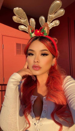 Woman with vibrant red hair wearing gold reindeer antlers and a white ribbed top, posing indoors against a red wall.
