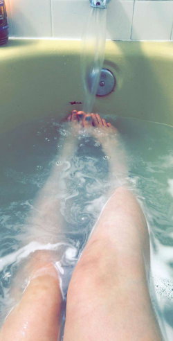 Person relaxing in a bubble bath with feet under running water, legs visible from knees down.