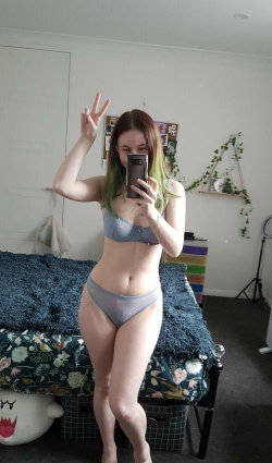 A woman with green-tipped brown hair takes a mirror selfie in a gray bra and matching thong, standing in front of a bed