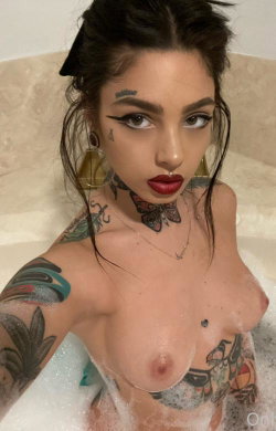 A tattooed woman with dark hair and bold makeup poses topless in a bubble bath, showcasing her curvy figure and neck