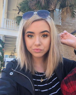 Blonde woman with shoulder-length hair, wearing sunglasses on head, black jacket over striped shirt, posing outdoors