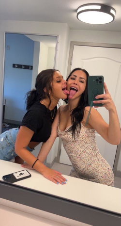 Two women posing playfully in a bathroom mirror selfie, one in a black crop top and denim shorts, the other in a floral