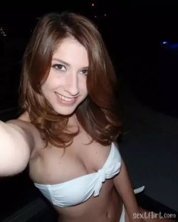 Brunette woman in a white bandeau top takes a selfie at night, smiling with shoulder-length hair and visible midriff.