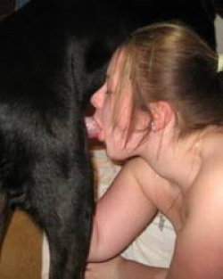 A woman with blonde hair is topless and engaging in oral contact with a large black dog indoors.