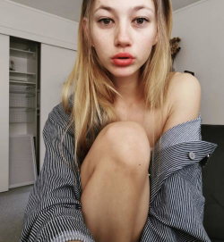 Blonde woman with shoulder-length hair, wearing an off-the-shoulder striped shirt, posing indoors with one knee raised.