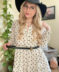 Blonde woman in a polka dot dress and black hat poses indoors with glasses and a belt, surrounded by plants.