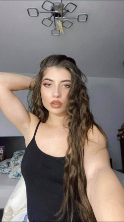Curvy woman with long dark wavy hair takes a selfie in a black tank top, posing in a bedroom with a bed and modern