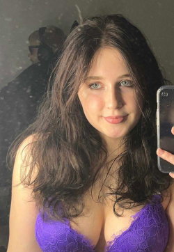 Woman with long dark hair and blue eyes wears a purple lace top, taking a mirror selfie in a dimly lit room.