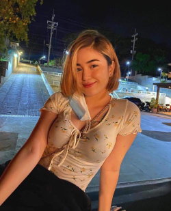A woman with short blonde hair in a floral top poses outdoors at night on a paved path, smiling at the camera.