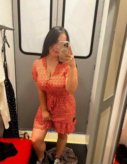 A woman with long dark hair takes a mirror selfie in a red polka-dot dress inside a fitting room, holding her phone