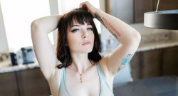 Brunette woman with tattoos on arms, wearing a light blue tank top, posing indoors with hands behind her head.