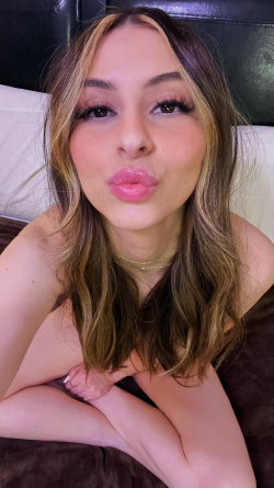A woman with long brown hair and highlights lies on a bed, making a playful kiss face toward the camera in a close-up