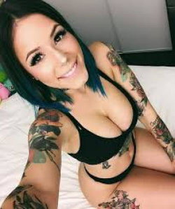 Brunette woman with blue-tipped hair and full arm tattoos poses in a black bra and thong on a bed, smiling at the