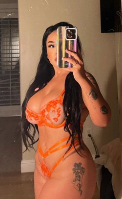 Curvy woman with long dark hair takes mirror selfie in orange lace lingerie, showing tattoos on arm and hip.