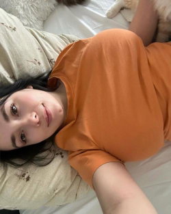 A woman with dark hair lies on a bed in an orange top, looking at the camera with a soft expression.