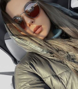 A woman with long brown hair and round sunglasses rests her head in a car, wearing a shiny olive puffer jacket.