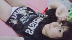 A young woman with short dark hair lies on a pink surface, wearing a black graphic tee and white underwear, looking at